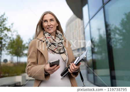 A businesswoman walks near a business center while holding a smartphone and documents A businesswoman walks near a business center while holding a smartphone and documents 119738795