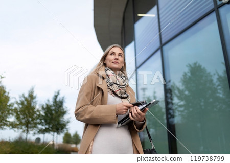 A businesswoman walks by a modern office building while holding a tablet and looking up A businesswoman walks by a modern office building while holding a tablet and looking up 119738799