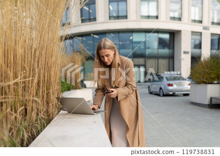 A businesswoman checks her phone while working on her laptop outside a business center 119738831