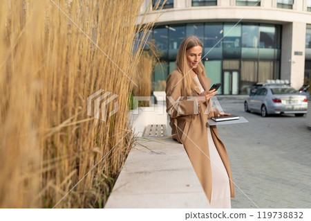 A businesswoman uses her phone while leaning against a wall outside a business center A businesswoman uses her phone while leaning against a wall outside a business center 119738832