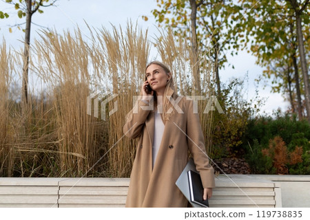 A businesswoman talks on her phone while holding a laptop near a business center A businesswoman talks on her phone while holding a laptop near a business center 119738835