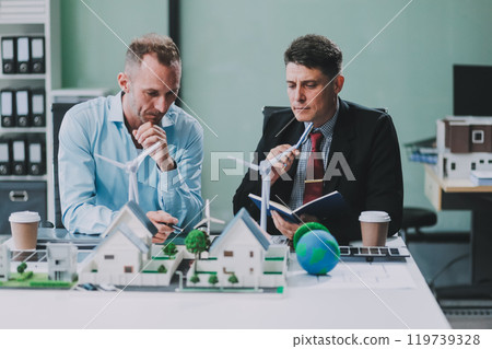 Engineers meeting about wind turbine model and house model. Renewable energy and housing development concept. Team of architects and engineers in modern office discussing design structure. 119739328