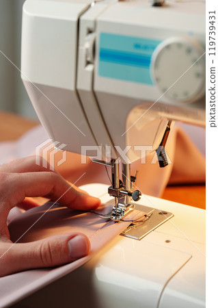 Hands sewing fabric using a sewing machine on a wooden table in a well-lit workspace 119739431