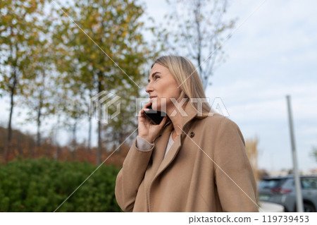 A businesswoman in a brown coat walks near a business center while talking on her phone A businesswoman in a brown coat walks near a business center while talking on her phone 119739453