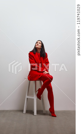 Woman dressed in red sitting on stool, looking upwards with calm and reflective expression. 119741029