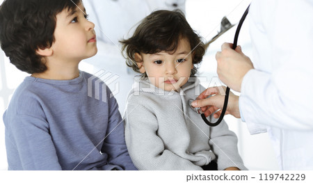 Woman-doctor examining a child patient by stethoscope. Cute arab toddler and his brother at physician appointment. Medicine concept Woman-doctor examining a child patient by stethoscope. Cute arab toddler and his brother at physician appointment. Medicine concept 119742229
