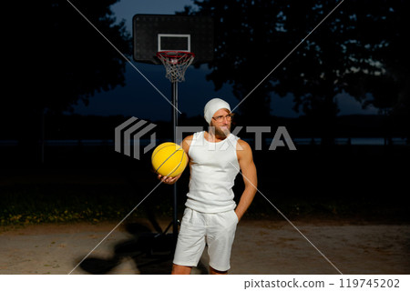 Man holding a basketball in a park at dusk, showcasing athleticism and confidence by the court under a darkening sky Man holding a basketball in a park at dusk, showcasing athleticism and confidence by the court under a darkening sky 119745202