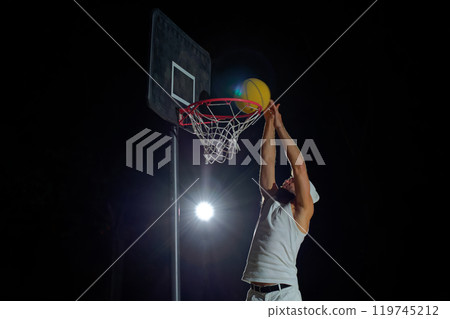 A man performs a slam dunk on a basketball court at night under the glare of bright outdoor lights 119745212