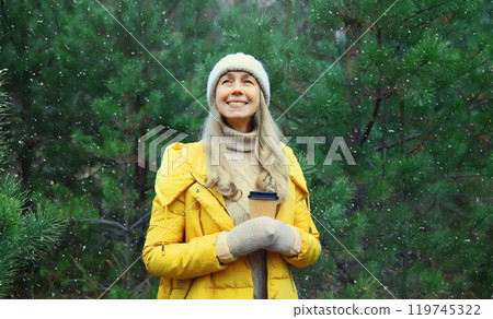 Winter day, happy smiling woman holding cup of coffee drink enjoys snow, warm weather against Christmas tree 119745322