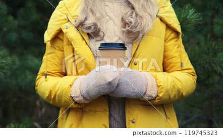 Close up female hand holding cup of hot coffee drink against Christmas tree in winter forest Close up female hand holding cup of hot coffee drink against Christmas tree in winter forest 119745323