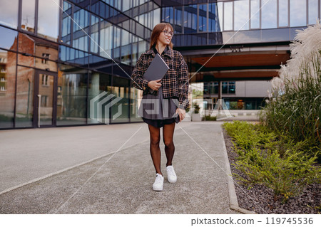 A Stylish Young Woman Walking on an Urban Campus while Carrying her Notebook, Embracing Life A Stylish Young Woman Walking on an Urban Campus while Carrying her Notebook, Embracing Life 119745536