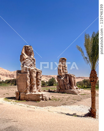 wo colossal statues of ancient Egyptian pharaohs stand in the vast desert landscape. The weathered statues and the surrounding ruins offer a glimpse into the rich history and mystery of ancient Egypt. 119746399