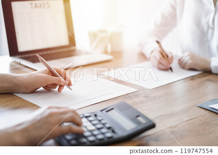 Two accountants using a laptop computer and calculator while counting taxes at wooden desk in office. Teamwork in business audit and finance 119747554