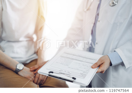 Doctor and patient in clinic. Friendly physician examining a young woman with a one hand while keeping a clipboard with medical records in another. Medicine concept Doctor and patient in clinic. Friendly physician examining a young woman with a one hand while keeping a clipboard with medical records in another. Medicine concept 119747564