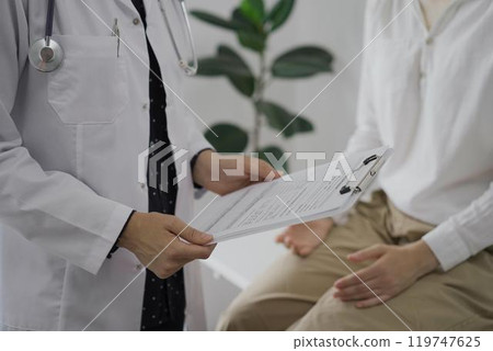 Doctor and a patient discussing something. The female physician, wearing a white medical coat is keeping clipboard with a medical notes besides a young woman during a consultation in the clinic 119747625