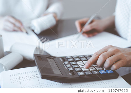 Two accountants use a calculator and laptop computer for counting taxes or revenue balance. Business, audit, and taxes concepts Two accountants use a calculator and laptop computer for counting taxes or revenue balance. Business, audit, and taxes concepts 119747633