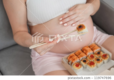 A pregnant woman sits on the sofa and eats rolls from a box. Food delivery. Close up of belly.  119747696