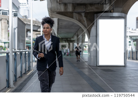 Teenage businesswoman, american african, afro hairstyle Holding a tablet walking on city street rushing to work. Empty white billboard for advertising. 119750544