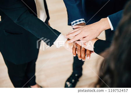 Colleagues from various ethnicities and backgrounds join hands in a modern office, emphasizing teamwork, cooperation, and community. An above view showcases their commitment to equality and diversity. 119752457
