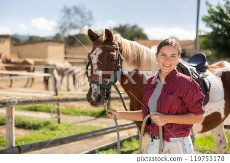 Happy young girl preparing horse for riding and walking in pasture on farm 119753178