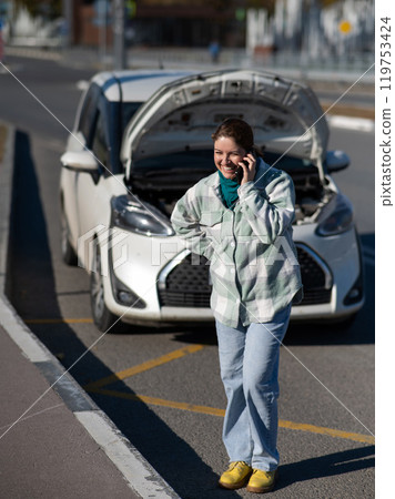 Caucasian woman laughing and calling on phone asking for help standing near car with open hood.  119753424