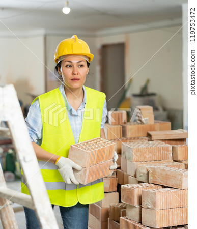 Workwoman in yellow vest and hardhat carrying hollow clay bricks on construction site indoors Workwoman in yellow vest and hardhat carrying hollow clay bricks on construction site indoors 119753472