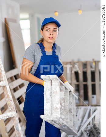 Woman builder in blue overalls carries a ladder in her hands in a room being renovated 119753563