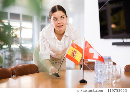 Positive young woman putting flag of Spain on the table with flag of Switzerland in conference room 119753623