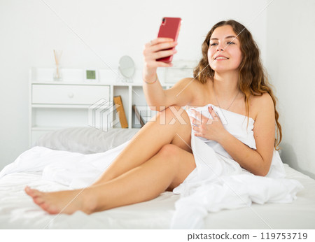 Naked girl covered with sheet taking selfie on bed 119753719