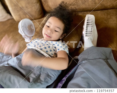 Little Asian girl playing with electric fan on sofa at home 119754357