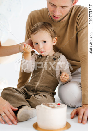 Father and son are sitting on the floor in front of birthday cake. Child boy is eating a piece of cake with a spoon. Scene is happy and playful Father and son are sitting on the floor in front of birthday cake. Child boy is eating a piece of cake with a spoon. Scene is happy and playful 119755867