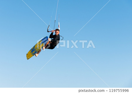 kiter performs difficult trick against blue sky background. sexy athletic man looks at camera cheekily while performing trick on kiteboard hanging in air 119755996