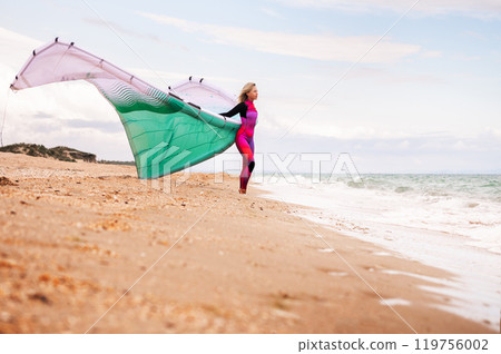 sportswoman in a wetsuit stands on a sandy beach between an inflatable kite and the foamy sea. The young woman enjoys the sport and the wind sportswoman in a wetsuit stands on a sandy beach between an inflatable kite and the foamy sea. The young woman enjoys the sport and the wind 119756002