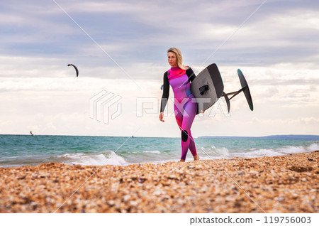 Young woman walking towards water on beach shore to do hydrofoil water sport. Sexy blonde girl in wetsuit walking along sea Young woman walking towards water on beach shore to do hydrofoil water sport. Sexy blonde girl in wetsuit walking along sea 119756003