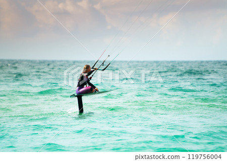 A young athlete in a colorful wetsuit sits on her hydrofoil and rides the waves with kitesurfing. Shot with a telephoto lens A young athlete in a colorful wetsuit sits on her hydrofoil and rides the waves with kitesurfing. Shot with a telephoto lens 119756004