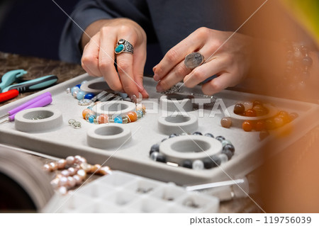 Top view artistic female hands creating bracelet from stones. POV shot of woman working with craft. Accessories on workshop table close up Top view artistic female hands creating bracelet from stones. POV shot of woman working with craft. Accessories on workshop table close up 119756039