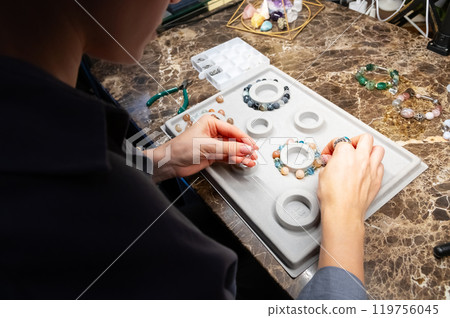 Top view artistic female hands creating bracelet from stones. POV shot of woman working with craft. Accessories on workshop table close up 119756045