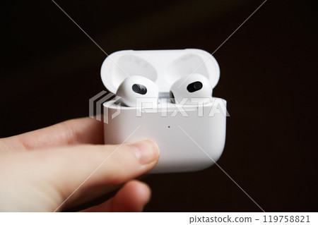 A close-up image of a hand holding wireless headphones in a sleek white case, set against a crisp black background A close-up image of a hand holding wireless headphones in a sleek white case, set against a crisp black background 119758821