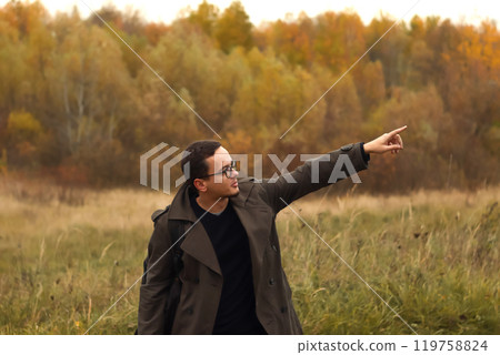 A young man in an autumn forest, pointing his finger to show direction. He is surrounded by vibrant fall foliage, with shades of orange, yellow, and red leaves creating a warm atmosphere A young man in an autumn forest, pointing his finger to show direction. He is surrounded by vibrant fall foliage, with shades of orange, yellow, and red leaves creating a warm atmosphere 119758824
