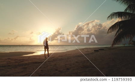 Woman is enjoying a beautiful sunset on the beach, with golden light reflecting off the ocean and palm trees swaying in the breeze 119759393