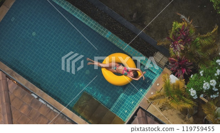 Young woman is enjoying a sunny day, floating on a yellow inflatable ring in a luxurious swimming pool surrounded by lush tropical vegetation 119759455