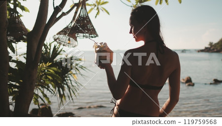 Young girl with coconut cocktail relax on a tropical beach. Palms, ocean, exotic landscape in sunset soft light. Travel, tourism, holiday in Thailand, Koh Phangan. Zoom in portrait, slow motion, 4K 119759568