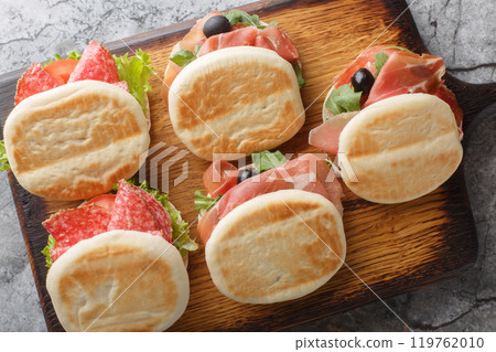 Small italian tigelle sandwiches with sausage, jamon and fresh vegetables close-up on wooden board. Horizontal top view Small italian tigelle sandwiches with sausage, jamon and fresh vegetables close-up on wooden board. Horizontal top view 119762010