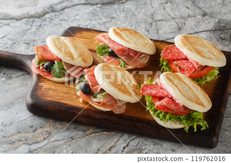 Delicious Italian Tigelle sandwich with salami, ham, olive, tomato, lettuce and arugula close-up on wooden board. Horizontal Delicious Italian Tigelle sandwich with salami, ham, olive, tomato, lettuce and arugula close-up on wooden board. Horizontal 119762016
