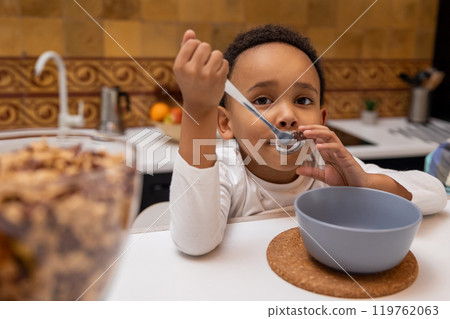 Dark-skinned boy sitting at the table and eating cereals Dark-skinned boy sitting at the table and eating cereals 119762063