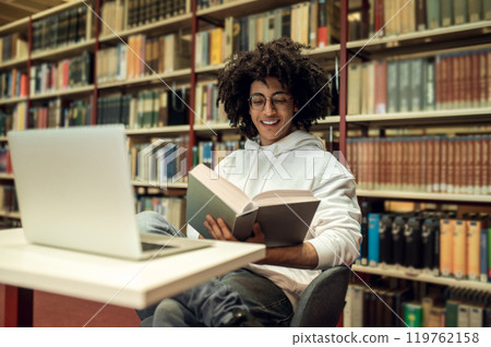 Curly-haired young man studying in the library and looking involved Curly-haired young man studying in the library and looking involved 119762158