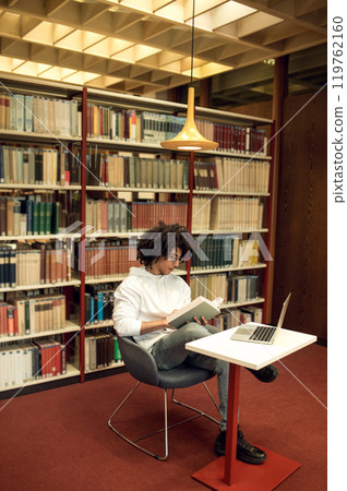 Curly-haired young man studying in the library and looking involved Curly-haired young man studying in the library and looking involved 119762160