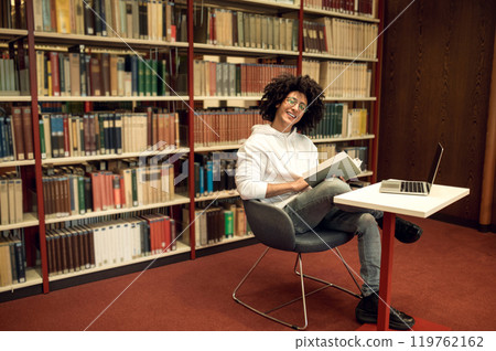 Curly-haired young man studying in the library and looking involved Curly-haired young man studying in the library and looking involved 119762162