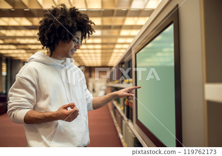 Curly-haired young man standing near the screen in the library and searching a book 119762173