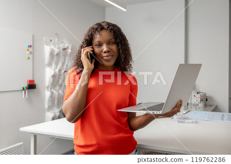 African american young seamstress with a laptop discussing something on the phone African american young seamstress with a laptop discussing something on the phone 119762286